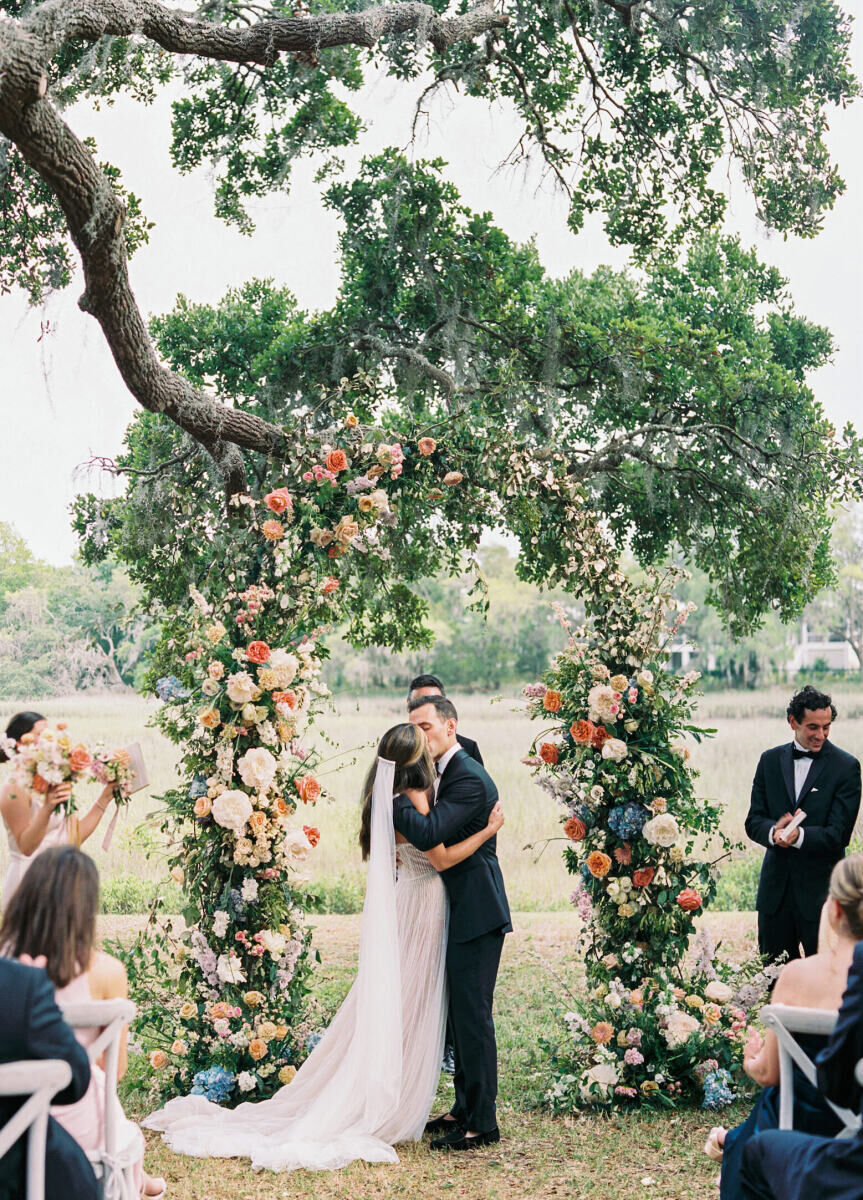 A bride and groom kiss in front of their colorful floral arch during their elegant spring wedding ceremony.